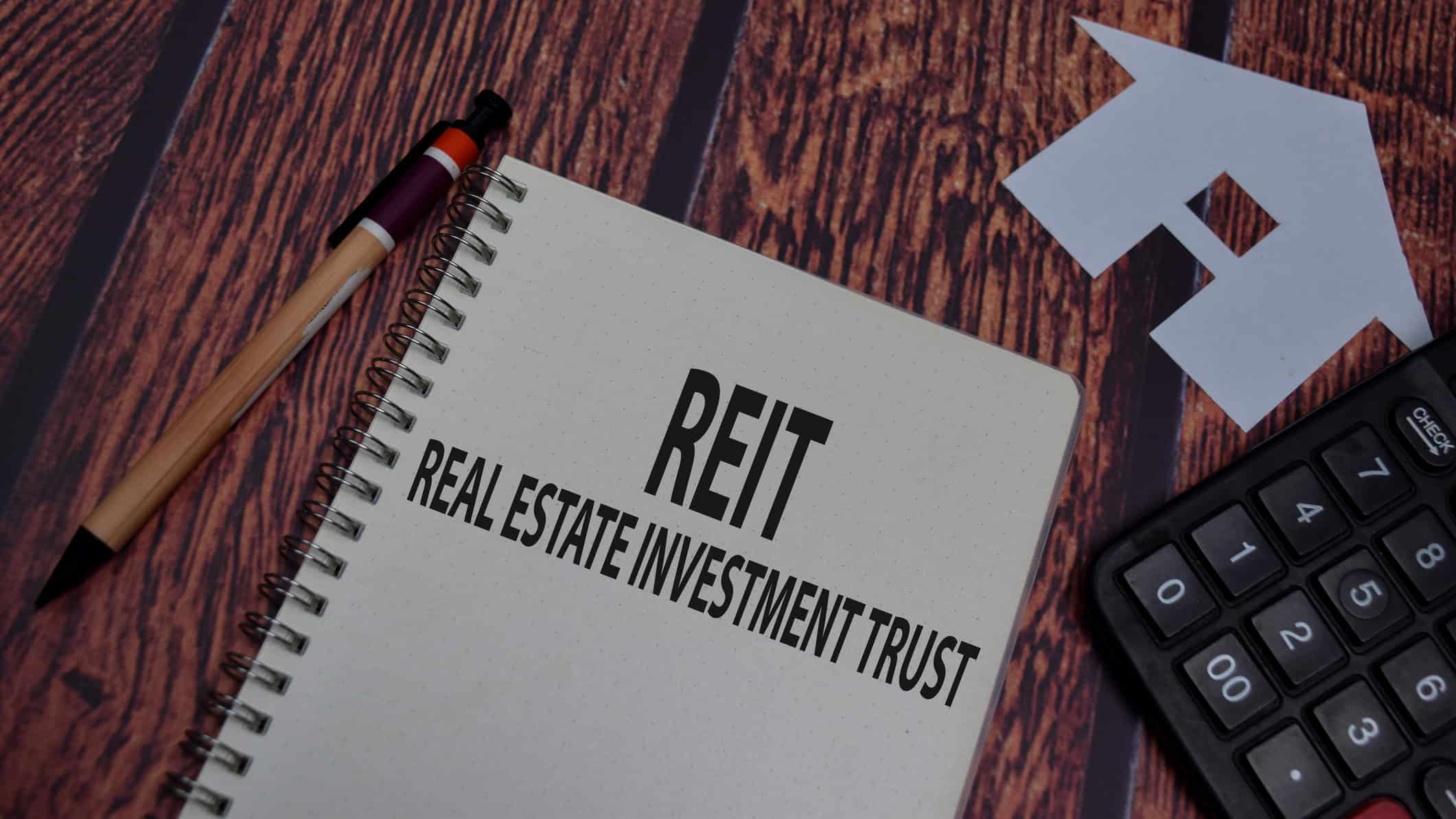 REIT and real estate investment trust written on a notebook with a pen, paper house, and calculator around it.