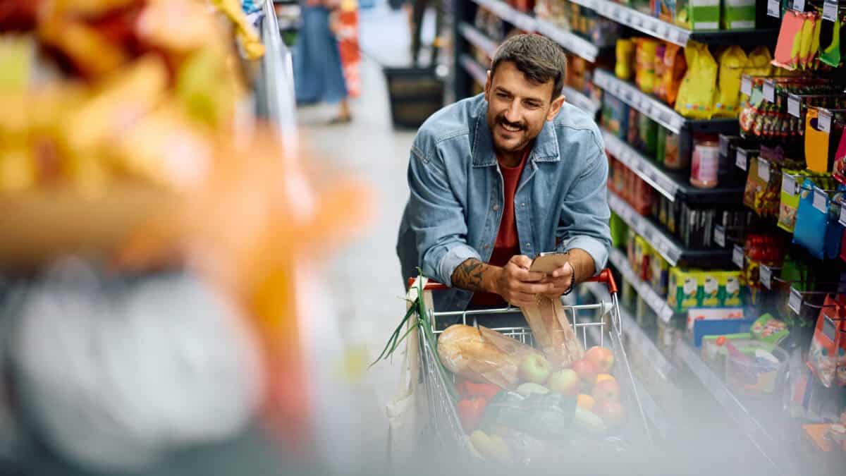 Smiling man with a full trolley looking at an aisle in the supermarket while holding his phone.