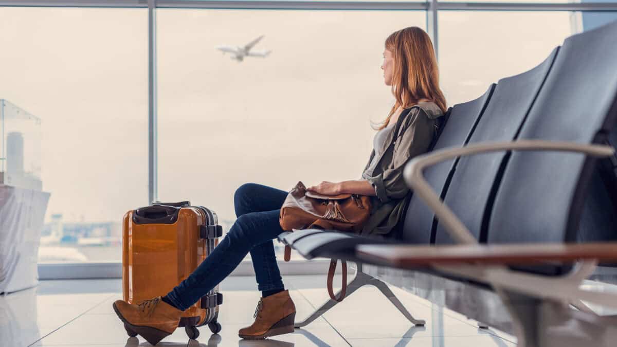 Woman looking out window at flying airplane while waiting to board in airport lounge.