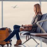 Woman looking out window at flying airplane while waiting to board in airport lounge.