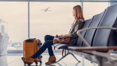 Woman looking out window at flying airplane while waiting to board in airport lounge.