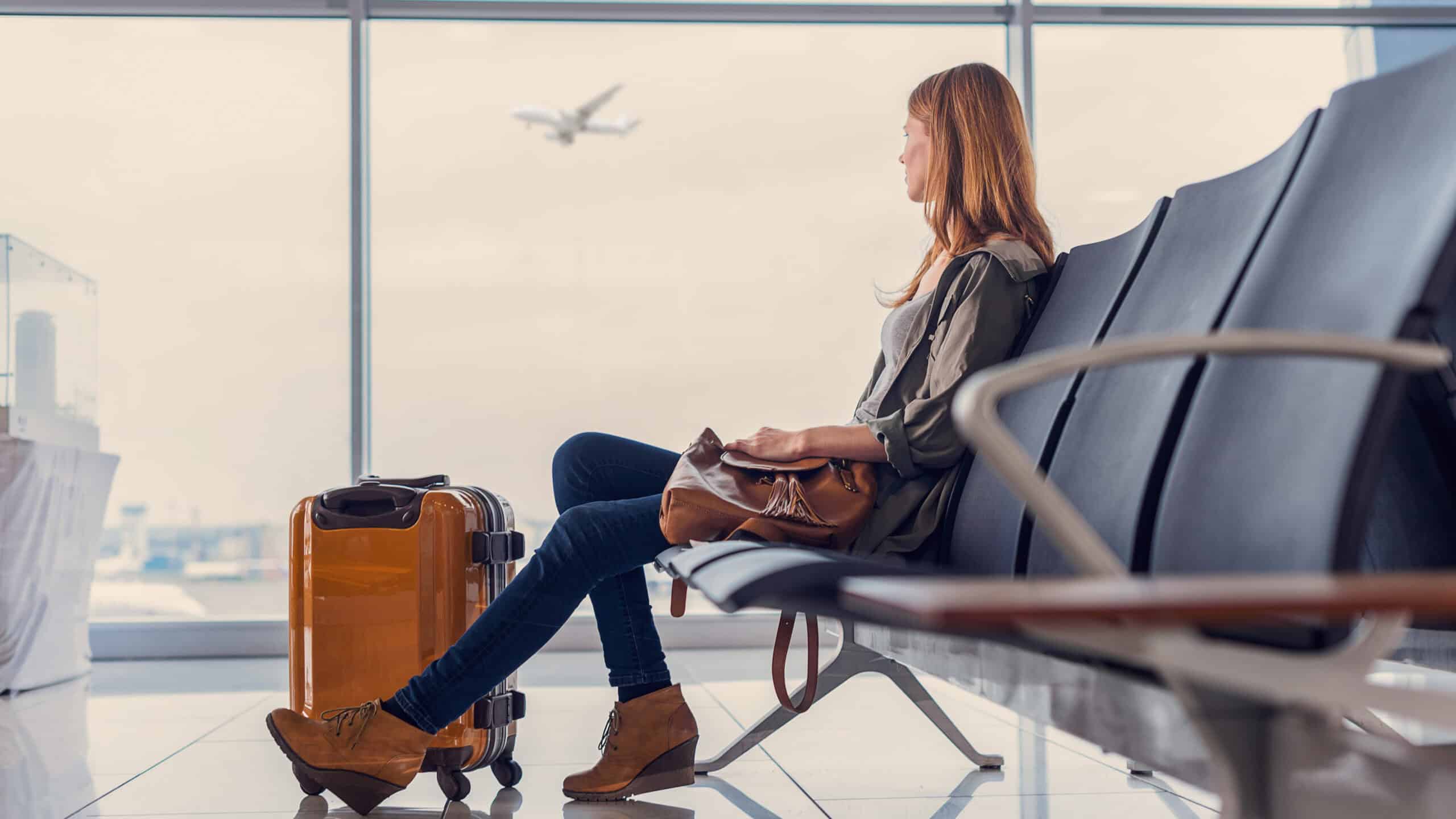 Woman looking out window at flying airplane while waiting to board in airport lounge.