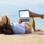 Man reading his emails on a laptop while relaxing on a beach.