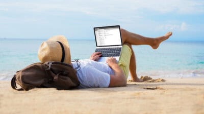 Man reading his emails on a laptop while relaxing on a beach.