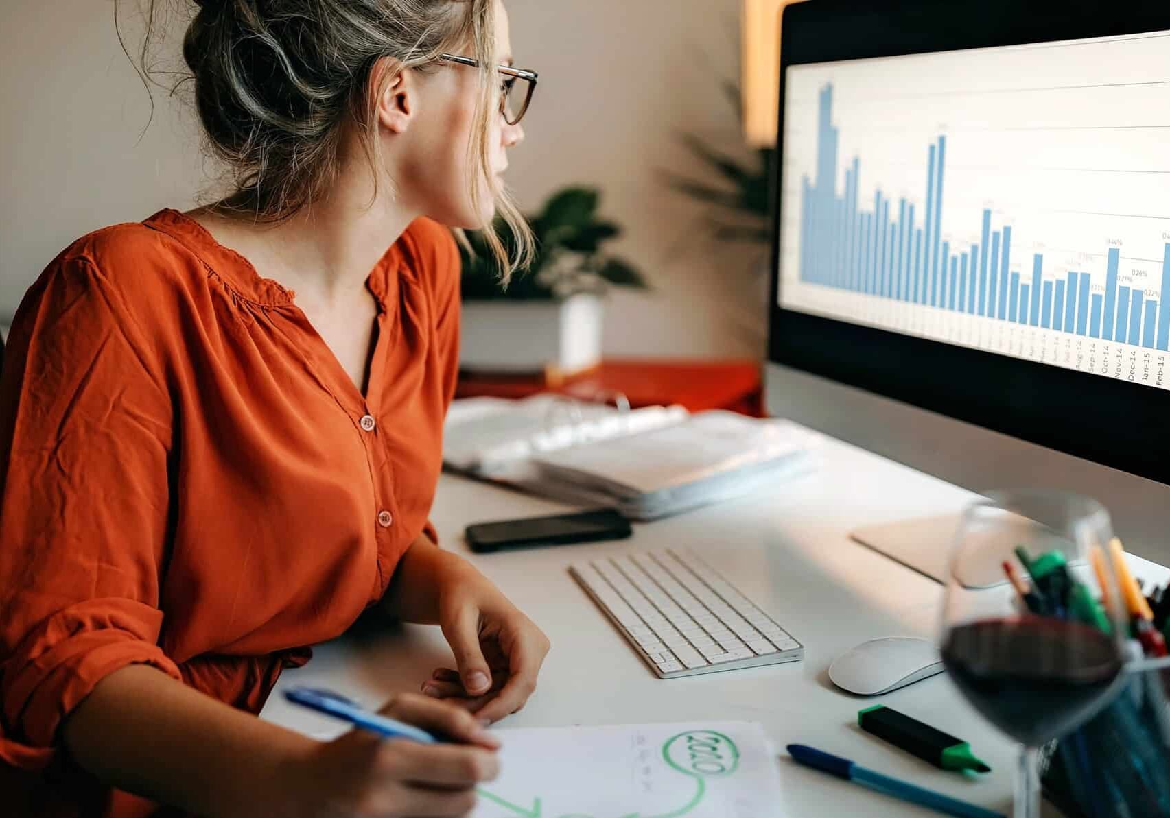A woman studying share market stats on a computer while writing a report.