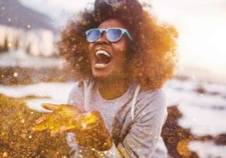 A woman blowing gold glitter out of her hands with a joyous smile on her face.